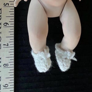 May include: A porcelain baby doll with white crocheted booties. The doll is sitting with its legs crossed and its feet are visible.