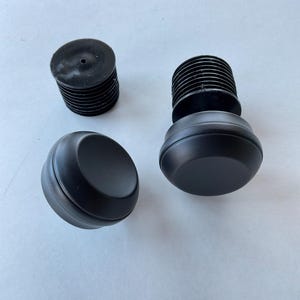 May include: Three black plastic parts for a furniture piece. Two of the parts are round and have a smooth, flat top. The third part is cylindrical with a ribbed texture.