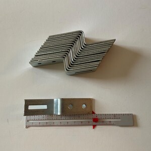 May include: A stack of 16 silver metal shelf brackets, each with two holes for mounting. A single bracket is shown next to a ruler, measuring 3 inches in length.
