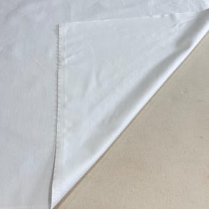 May include: A folded white fabric, possibly a sheet or tablecloth, with a visible seam along one edge. The fabric is smooth and appears to be made of a lightweight material. The corner is folded over, revealing the underside of the fabric.