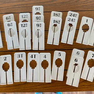 May include: White plastic garment size labels with black text. The labels are for sizes 3, 6, 9, 12, 18, 24, 1, 1T, 2, 2T, 3T, 4T, 4, 5, 6, and 7.