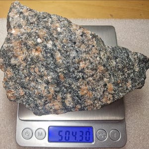 Op de afbeelding: Een ruw, onregelmatig stuk graniet met een gespikkelde textuur van zwarte, witte en oranje-roze minerale korrels. De steen rust op een digitale weegschaal die een gewicht van 504,30 gram weergeeft op een helder blauw verlicht scherm.
