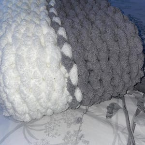 May include: A white and grey knitted throw pillow with a fluffy texture. The pillow is made of yarn and has a soft, cosy feel.
