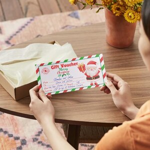 Puede incluir: Un vale de regalo rojo y verde con un Pap&aacute; Noel de dibujos animados sosteniendo un regalo. El vale dice "Vale de regalo Feliz Navidad" y tiene un espacio para el nombre del destinatario y una lista de regalos.