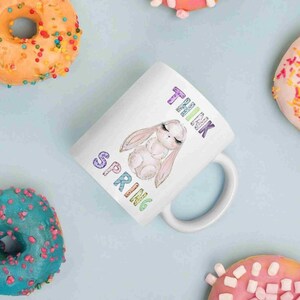 May include: White ceramic mug with a rainbow colored "Think Spring" text and a cartoon bunny with closed eyes. The mug is surrounded by donuts with colorful sprinkles.