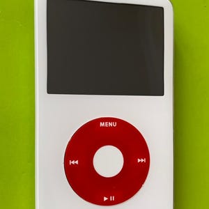 以下が含まれることがあります： 「MENU」と書いてある赤い丸いボタンが付いた白いiPod。ボタンの中央には白い円があり、円の両側には白いボタンがあります。iPodは黒い画面です。