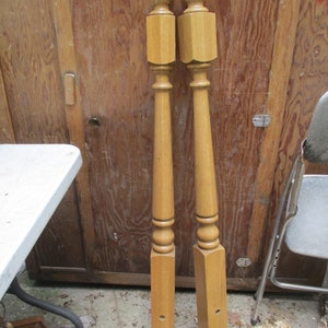 May include: Two wooden balusters with a light brown finish. Each baluster has a rounded top, a square midsection, and a long, tapered body. The bottom section is rectangular with pre-drilled holes. They are leaning against a wooden door.