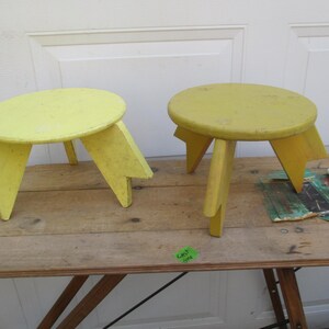 May include: Two small, round, yellow wooden stools with three angled legs each. The stools are painted in a faded yellow color and sit on a wooden table. The stools are likely vintage or antique.