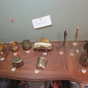 May include: A collection of nine vintage gold-toned metal items, including a small basket, a bell, a small pitcher, and several decorative bowls. The items are arranged on a wooden surface with a handwritten sign that reads "Choice".