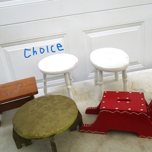 May include: Four wooden stools, one with a red finish and a white painted design, one with a brown finish and a white painted design, and two with a white finish. The stools are arranged on a white surface. The word "Choice" is written in blue on the white surface.