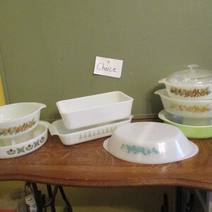 May include: A collection of vintage Pyrex dishes in various shapes and sizes. Some dishes feature floral patterns in orange, yellow, and green. Other dishes are plain white or light green. The word "Choice" is written on a small piece of paper.