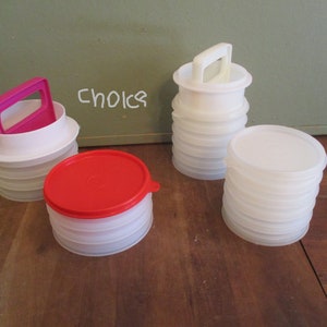 May include: Several white plastic containers with lids and handles are displayed. One container has a red lid, and another has a pink handle. The word "Choice" is written on the wall in the background. The containers appear to be for food storage.