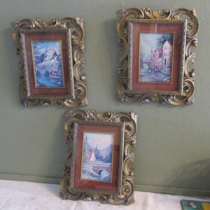 May include: Three framed landscape paintings with ornate gold-colored frames. Each painting features a different scene: a mountain range, a European village with a bridge, and a snow-covered mountain with a red-roofed house.