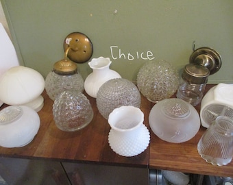 Może przedstawiać: Kolekcja 10 vintage'owych kloszy lamp wykonanych ze szkła w różnych stylach i kolorach, w tym przezroczystych, matowych i białych. Niektóre klosze mają teksturę lub wzór. Klosze są ułożone na drewnianej powierzchni.