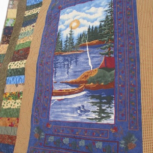 May include: A blue quilted fabric with a scene of a lake with trees and a canoe. The lake is surrounded by a brown border with a checkered pattern.