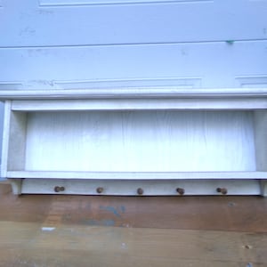 May include: A white wooden shelf with a small ledge and four wooden pegs below. The shelf is painted in a light, off-white color and appears to be handmade. The shelf is designed for storage and display.