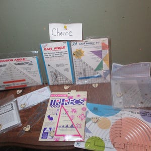 May include: A collection of quilting tools and rulers, including a "Companion Angle" ruler, two "Easy Angle" rulers, a "Tri-Recs Tools" ruler, and a "Easy Circle Cut" ruler. The rulers are on a wooden surface with a power strip and a few small heart-shaped objects.