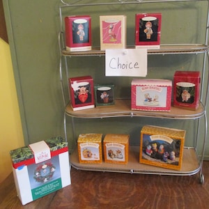 May include: A three-tiered metal shelf displays various boxed ornaments. The boxes are red, white, and orange, with illustrations of figures and scenes. One box features a wreath ornament. The word "Choice" is written on a white paper.
