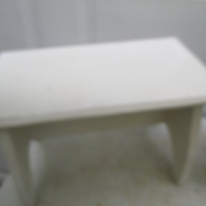 May include: A small, rectangular, white wooden step stool. The stool has a flat top and four angled legs. The design is simple and functional, suitable for various uses around the home. The stool is painted white.