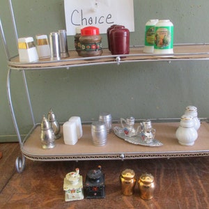 May include: A two-tiered metal shelf displays a collection of vintage salt and pepper shakers, spice containers, and a small tea set. The items are in various materials, including ceramic, metal, and glass. The sign says "Choice".