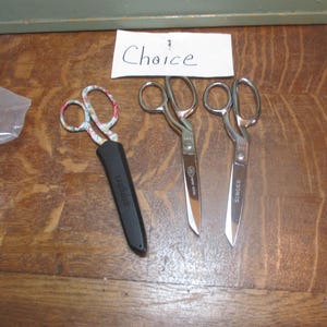 May include: Three pairs of scissors on a wooden surface. The scissors on the left have a floral pattern on the handles and a black plastic sheath. The two scissors on the right are silver with black handles and the words "SINGER" printed on them. The word "Choice" is written on a white card above the scissors.