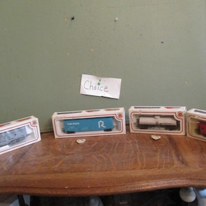 May include: Four model train cars in their original boxes, displayed on a wooden surface. The cars are various colors, including red, blue, and white. The boxes have a red and white design.