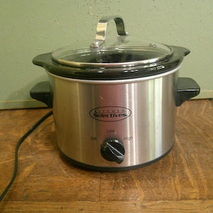 May include: A stainless steel Kitchen Selectives slow cooker with a clear glass lid and a black plastic handle. The front features a black control knob with settings for Off, Low, and High, along with a black power cord extending to the left side.