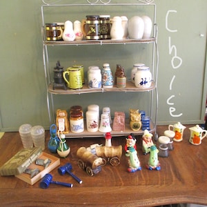 May include: A collection of vintage salt and pepper shakers and other decorative items displayed on a three-tiered shelf and a wooden table. The collection includes various shapes, colors, and designs, such as miniature mugs, bottles, and figurines.