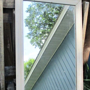 May include: Rectangular mirror with a cream-colored wooden frame. The mirror reflects a view of a blue-green building with a white trim and a tree with green leaves against a blue sky. The mirror is leaning against a wooden surface.