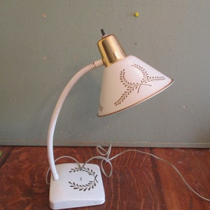 May include: Vintage white desk lamp with a gooseneck arm and a conical shade. The shade features a gold rim and a decorative leaf pattern. The base is white and square, with a matching leaf design. The lamp's top is gold.