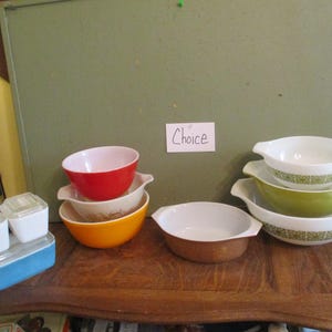 May include: A collection of vintage Pyrex bowls in various colors, including red, orange, and green, with white interiors and decorative patterns. Also, a set of white and blue plastic containers with clear lids are visible.