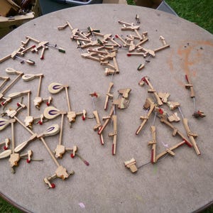 May include: A collection of disassembled piano parts, including hammers, dampers, and action components, scattered on a round, light brown table. The wooden parts have red, white, and purple accents. The scene suggests piano repair or restoration.