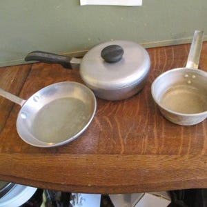 May include: Three silver aluminum cookware pieces: a small saucepan with a lid, a small frying pan, and a small sauce pot. The cookware is on a wooden surface.