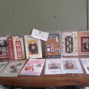 May include: A collection of 11 cross stitch patterns featuring various designs, including a heart trio, a coffee cup, a garland, a sweater, a  'Holly Decor' design, a 'Simply Designs' design, a 'Sewing Basket' design, a 'Coffee Day Garden' design, a 'Monogram Bag' design, and a 'Simply Designs' design. The patterns are printed on white paper with black text and images.