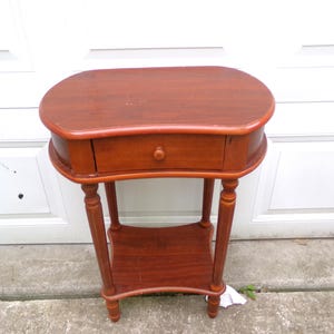May include: A kidney-shaped wooden side table with a warm brown finish, featuring a single central drawer with a small round knob, four slender turned legs, and a lower shelf for storage. The tabletop has a smooth, curved edge design.