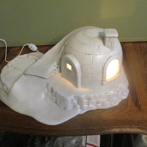 May include: A white ceramic igloo-shaped lamp with a chimney and two arched doorways. The lamp is lit from within, casting a warm glow. The lamp sits on a base that resembles a snowy landscape.