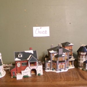 May include: A collection of miniature ceramic houses with intricate details. The houses feature various architectural styles, including red brick and white trim. Some have snow-covered roofs and decorative elements. A sign that says "Choice" is in the background.