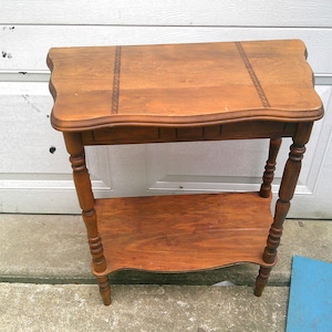 May include: A medium-brown wooden side table featuring a rectangular top with scalloped edges and two decorative inlaid strips. The table stands on four turned legs and includes a lower shelf with a matching scalloped edge design.
