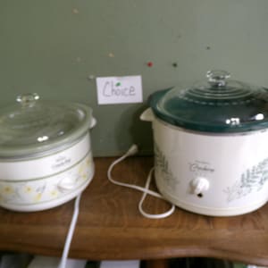 May include: Two white electric slow cookers with glass lids and floral patterns. The left unit features yellow flowers and the text Crock-Pot, while the right unit has green foliage and the text Crockery. A small white sign between them reads Choice.