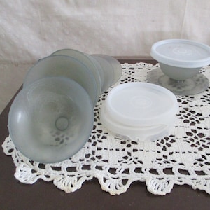 May include: A collection of vintage, light blue glass dessert dishes with matching white plastic lids. The dishes are stacked and arranged on a white lace doily. One dish is assembled with a lid, and another set of lids is stacked nearby.