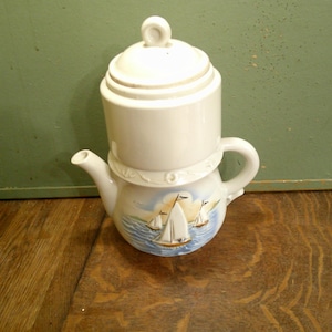 May include: A white ceramic teapot with a tall, cylindrical lid and a rounded base. The body of the teapot features a painted scene of sailboats on blue water with a soft yellow sky. It includes a curved handle and a small, pointed spout.