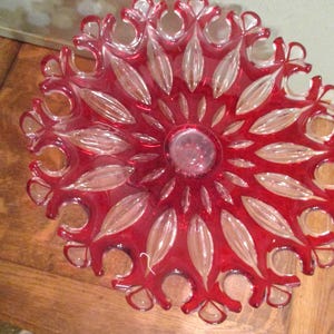 May include: A decorative red glass bowl with a floral design. The bowl features a central circular area surrounded by elongated, petal-like shapes. The rim has a scalloped edge. The bowl is likely vintage or antique.