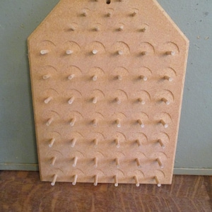May include: A light brown wooden pegboard with a hexagonal top. The board has rows of small wooden pegs and curved cutouts. A small hole is at the top center. The pegboard is likely for organizing items.