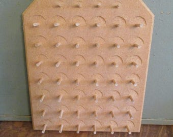 May include: A light brown wooden pegboard with a hexagonal top. The board has rows of small wooden pegs and curved cutouts. A small hole is at the top center. The pegboard is likely for organizing items.