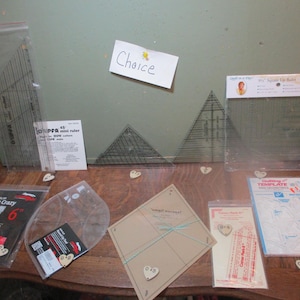 May include: A collection of quilting tools and templates, including a 45 degree mini ruler, a 6 inch scrap crazy ruler, a 12 inch square up ruler, a quilting template, and a variety of clear plastic rulers. The tools are arranged on a wooden surface with small heart-shaped decorations.