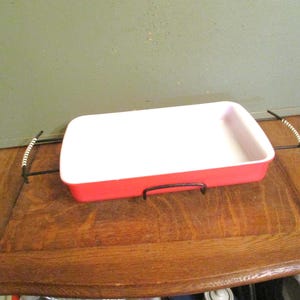 May include: A rectangular baking dish with a white interior and a red exterior. The dish is supported by a black metal frame with handles. The handles have a white and black twisted design. A kitchenware item.