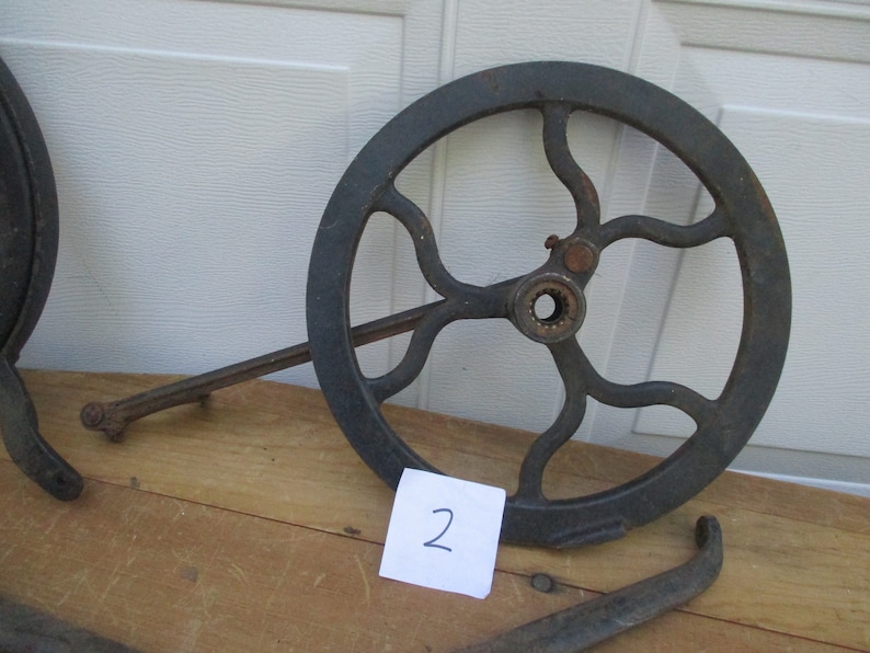 May include: A dark gray, cast iron wheel with a series of curved spokes and a central hub. A long, thin arm extends from the wheel's edge. A small piece of paper with the number "2" is visible.