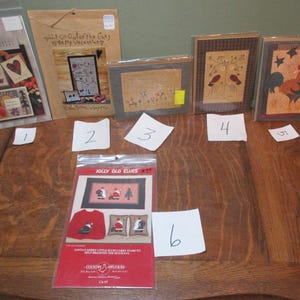 May include: Six different cross-stitch patterns on a wooden table. The patterns are for a variety of themes, including "Forever Friends", "Jolly Old Elves", "Don't Go Out of the Lines Be My Valentine", and "Santa's Merry Little Elves Can't Stay at Home Without Him for the Holidays".