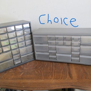 May include: Two plastic storage organizers with multiple small drawers. One is dark gray, the other is light gray. The word "Choice" is written in blue marker on the wall behind the organizers. These organizers are ideal for storing small items.
