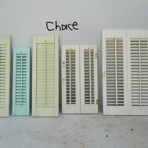 May include: Assortment of vintage wooden shutters in various sizes and colors. The shutters are painted in shades of cream, light green, and blue. The word "Chace" is written in black marker on the wall above the shutters.